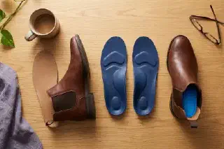 Overhead flat-lay of orthotic insoles next to a Chelsea boot with its factory insole removed on a warm wood desk surface