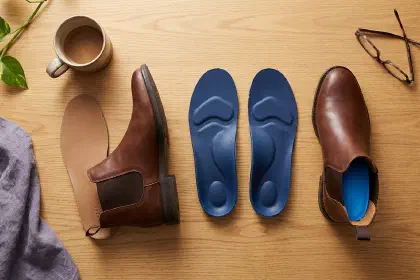Overhead flat-lay of orthotic insoles next to a Chelsea boot with its factory insole removed on a warm wood desk surface