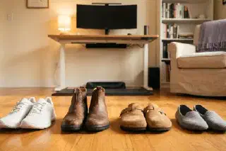 Four types of standing desk footwear in a row on hardwood — cushioned trainer, Chelsea boot, cork clog, and flat slipper — with a standing desk in soft focus behind