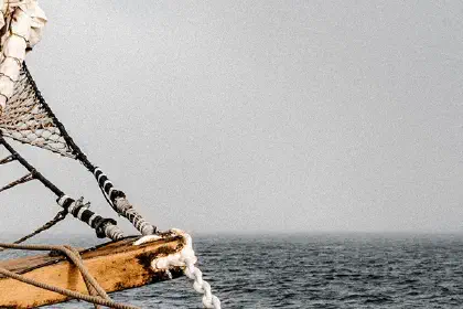 Wooden ship bow with rope rigging sailing across a gray open sea