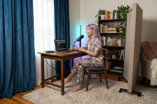 Mira recording at a small apartment desk with curtains, rug, bookshelf, and a movable acoustic panel