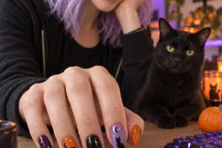 Mira with purple hair and black-cat Halloween nails at a desk lit by purple and warm orange light, a black cat and a small pumpkin beside a backlit keyboard